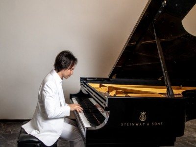 Piano de cola sobre un escenario con iluminación cálida y filas de butacas vacías.
